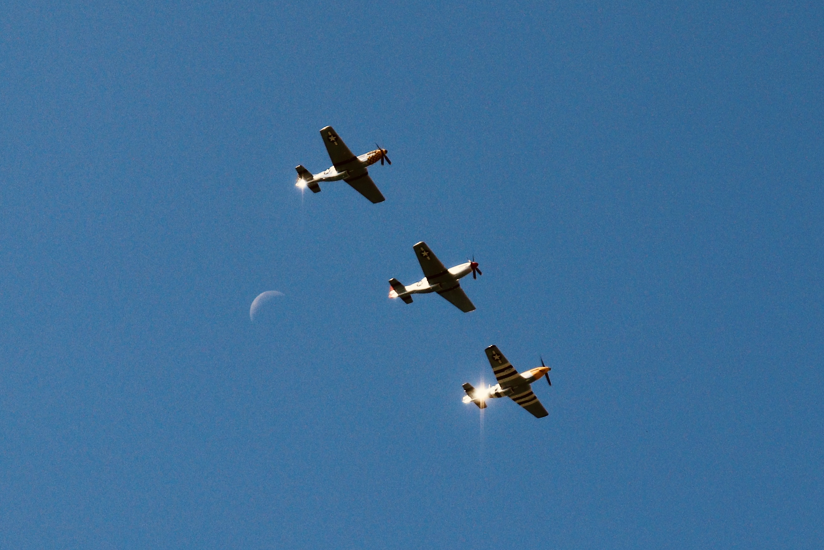 Three P-51 Mustang fighter planes in tight echelon formation banking across azure sky with crescent moon visible